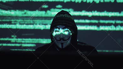 A male robber in a mask works on a computer in a dark room. computer code is reflected on his face. the hacker breaks