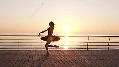 Beautiful silhouette of ballerina in ballet tutu and point on embankment above ocean or sea at sunrise. Young beautiful