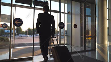 Unrecognizable man walking through glass automatic door of modern airport to city street and pulling suitcase on wheels