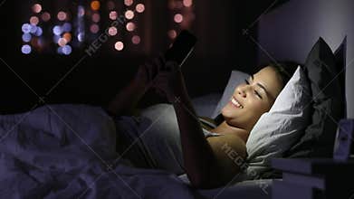 Happy girl chatting in a phone in the bed in the night