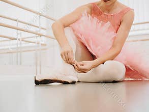 Young ballerina in pink tutu costume wraps white silk ribbons of soft top ballet shoes pointe and ties them up. Woman