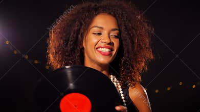 African-american young woman in party dress holding vinyl record and dancing on black lights background. Girl smiling