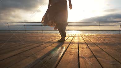 Young ballerina in beige silk dress running in pointe shoes on wooden embankment to sea or ocean. Sunrise background