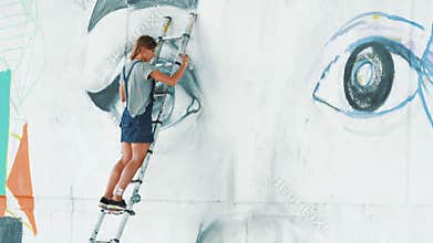 Beautiful girl making graffiti of big female face with aerosol spray on urban street wall. She standing on ladder
