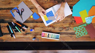 Zoom-out of creative brunette making paper collage drawing with crayons sitting at desk in home office. Artists