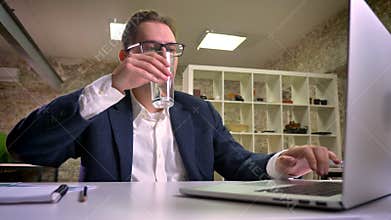 Close-up caucasian business guy in black suit printing on his laptop and drinking water while sitting at his workplace