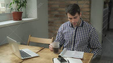 Attractive man drinking coffee and reading the newspaper, is on the table phone and a laptop.