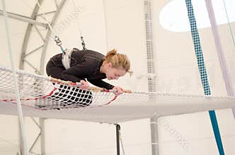 An adult female lands on a net, preparing to dismount at a on a flying trapeze school at an indoor gym. The woman is an amateur