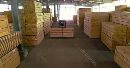 A large warehouse of timber materials. A forklift carries a wooden beam through the warehouse. Neatly stacked wooden bar
