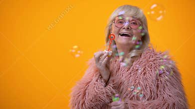 Funny stylish granny in pink coat and round sunglasses making soap bubbles, ad