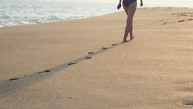 Rear view of female feet walking on golden sand at the beach with ocean waves at background. Legs of young woman