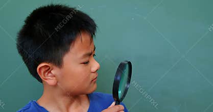 Front view of Asian schoolboy looking through magnifying glass in classroom at school 4k