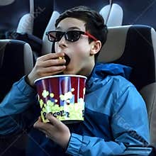 Guy teen in 3d glasses sits in a cinema chair and eats popcorn. The emotion of surprise on the boy`s face while watching an