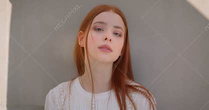 Pretty young ginger model flirting with camera leaning on the white wall.