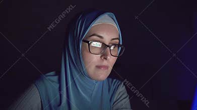 Muslim woman`s face in hijab and glasses at night in the dark uses computer