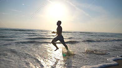 Silhouette of young sporty man running fast along sea shore during sunrise. Athletic boy training barefoot on the beach