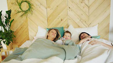 Young funny boy wake up while his parents sleep in morning on bed in their house