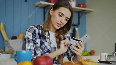 Smiling woman doing online shopping using smartphone and credit card while have breakfast in the kitchen at home