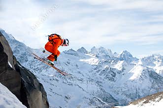 A professional skier makes a jump-drop from a high cliff against a blue sky leaving a trail of snow powder in the