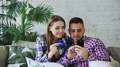Attractive young couple with smartphone and credit card shopping on the internet sit on couch in living room at home