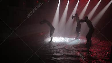 A group of guys are dancing active dance in water with splashes in the beam of a spotlight