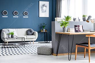 Dark blue living room interior with three clocks, simple poster, bright sofa and home office corner with laptop on hairpin desk