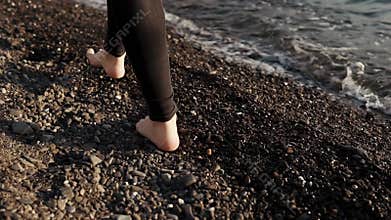Barefoot legs of slim young woman are stepping over pebble beach near seawater