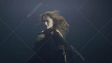 Mysterious woman in black dress swimming like mermaid under water in dark pool
