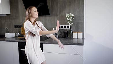 Alone crazy brunette woman is wearing pajamas and dancing in kitchen in evening, shaking her hands and body