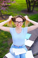 Smiling female student reading papers and playing with hair in spring park.