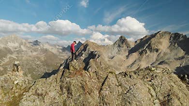 A young hipster guy with a beard a climber in a cap and sunglasses is standing on a rocky ridge high in the mountains