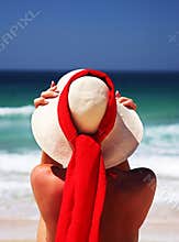 Girl sitting on sandy beach in the sun adjusting hat. Blue sky, blue sea red scarf. Spain.