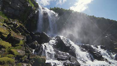 Beautiful view of the waterfall. High cliffs covered with green moss, the sun shines in the frame, water drops fall on
