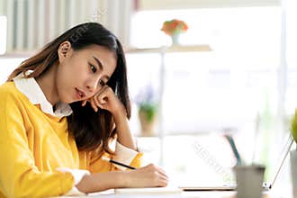 Young attractive asian female student sitting at table thinking and writing journal by hand note idea script, diary or sketch desi