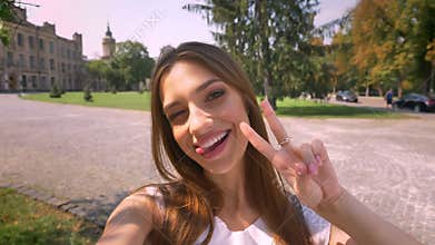 Nice young woman is standing in park in daytime, holds camera, makes selfie, show peace sign, communication concept