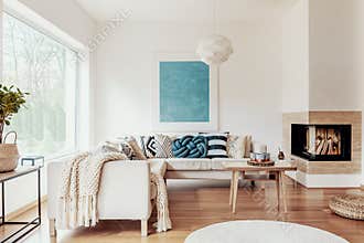 Turquoise blue knot pillow on a beige corner sofa and an abstract poster on a white wall in a modern living room interior