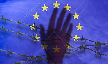 The shadow of the hand behind the barbed wire on the background of the EU flag.Metal fence with barbed wire on a European Union