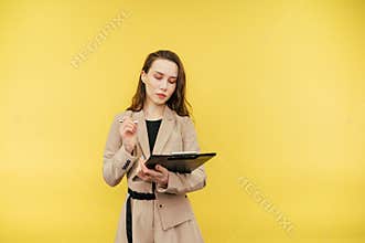 Business woman in a jacket stands on a yellow background and writes with a pen on a tablet with a serious face, isolated