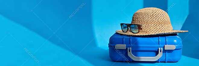 Suitcase straw hat and sunglasses in bright blue studio baner