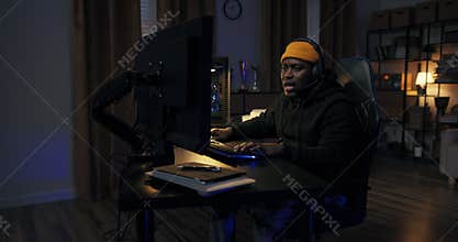 A man's success in a computer game. Boy in sweatshirt cap with headset raises hands up showing strength, victory