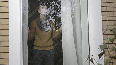 Young Caucasian brunette man standing behind window at home and closing curtains. Portrait of sad lonely guy spending