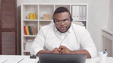Workplace of freelancer. African-American man works at home office using computer and headset. Employee is having a