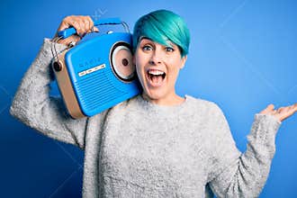 Young woman with blue fashion hair listening to music holding vintage portable radio very happy and excited, winner expression