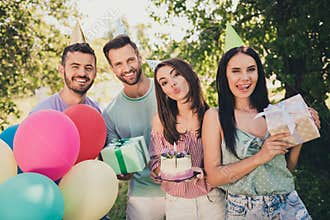 Photo of joyful brown haired happy friends at birthday party wear cones hold gift box cake balloons outside in picnic
