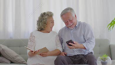 Happy Wellness Asian Elderly or Seniors couple holding mobile phone and book smile and laugh together on sofa in living room at