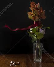 A red ribbon is moved by the wind around a glass vase containing flowers and leaves on a wooden table with vintage scissors