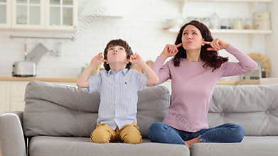 Frowning mom and son sit on couch with closed eyes and cover ears from noisy music or fight sounds