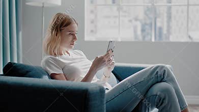 Blonde woman surfs internet with phone sitting on azure sofa