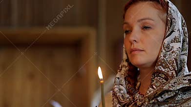 a Russian woman in a headscarf holds a candle in her hands and stands in front of icons in an Orthodox church and prays