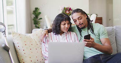 Happy biracial couple embracing in living room using laptop and smartphone, crutch leaning on couch
